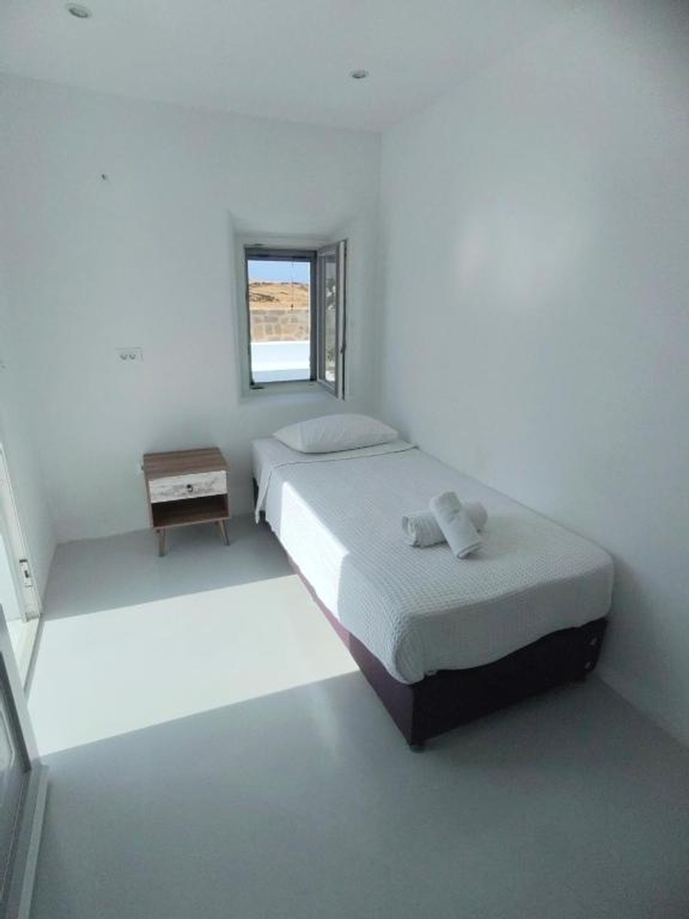 A minimalist bedroom with a single bed, a nightstand, a window with a view, and a small wooden cabinet.
