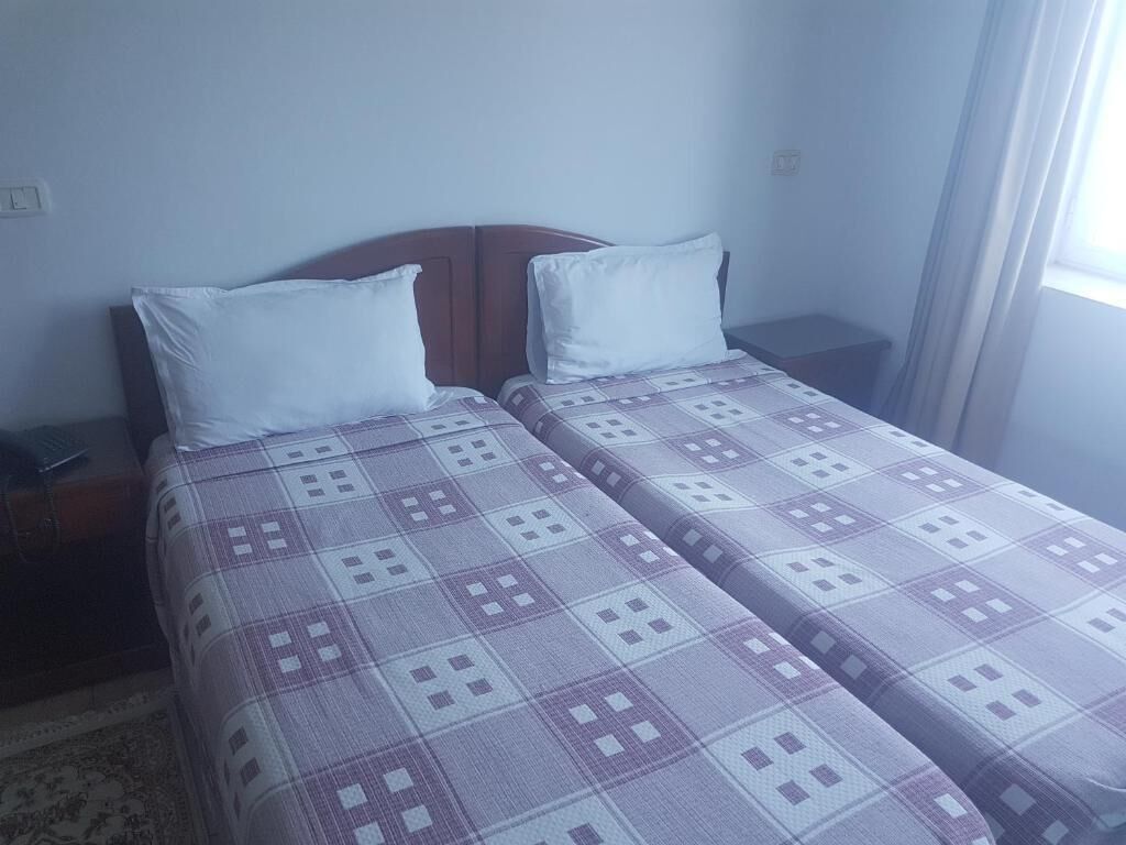 Een tweepersoonsbed met een geruite sprei, een houten hoofdbord en een nachtkastje met een lamp.