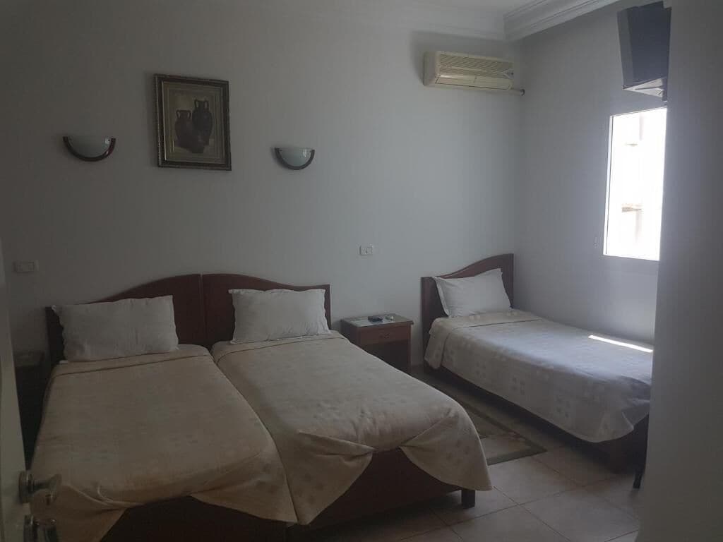 A room with two beds, a wall-mounted air conditioner, a small table, and a window with natural light.