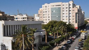 Un edificio de varios pisos con un diseño arquitectónico moderno, un estacionamiento con coches y palmeras bordeando la calle.