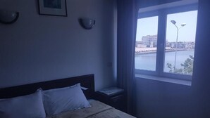 Un dormitorio con cama, mesita de noche, ventana con vista al agua y edificios, e iluminación en la pared.