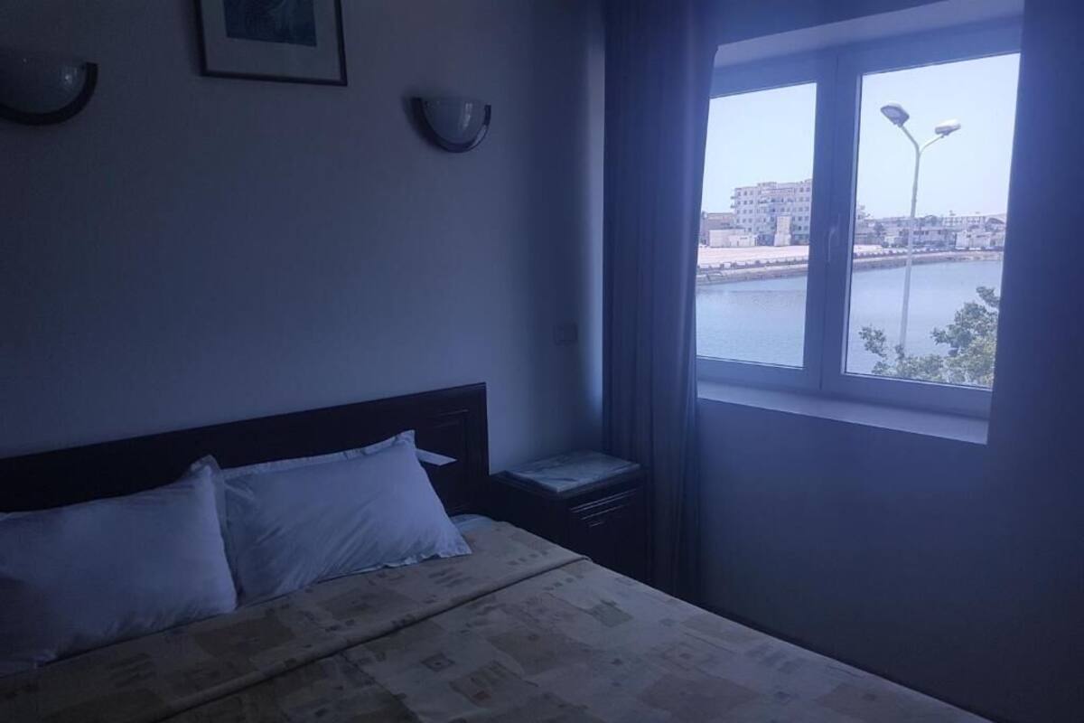 Un dormitorio con cama, mesita de noche, ventana con vista al agua y edificios, e iluminación en la pared.