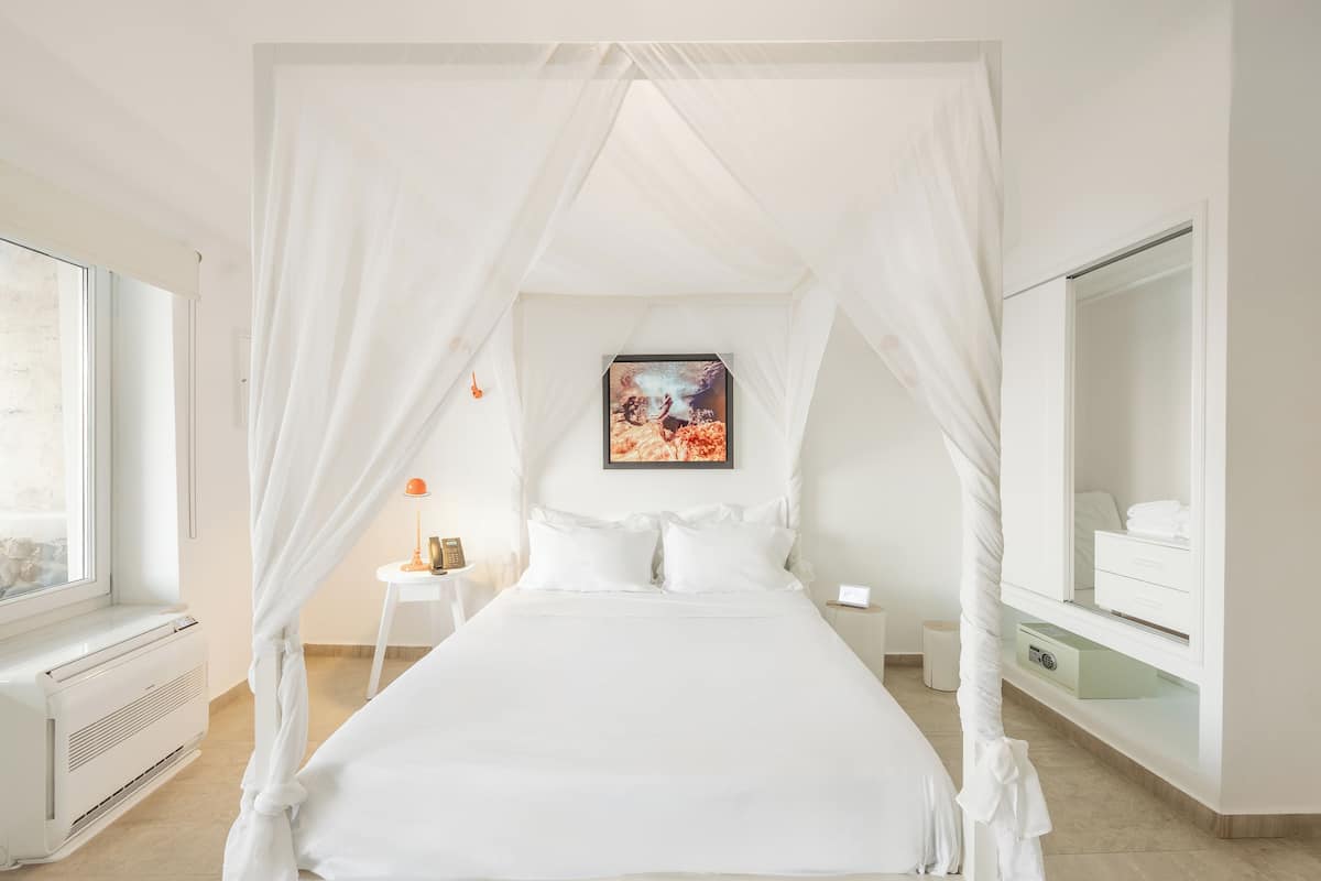 Ein Bett mit Baldachin, ausgestattet mit weißer Bettwäsche, ein Nachttisch mit Lampe, ein kleinerer Nachttisch mit einer Uhr und ein großer Spiegel auf der rechten Seite.
