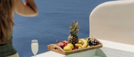 A person in a white bathrobe sitting by a bathtub with a fruit tray and a glass of water.