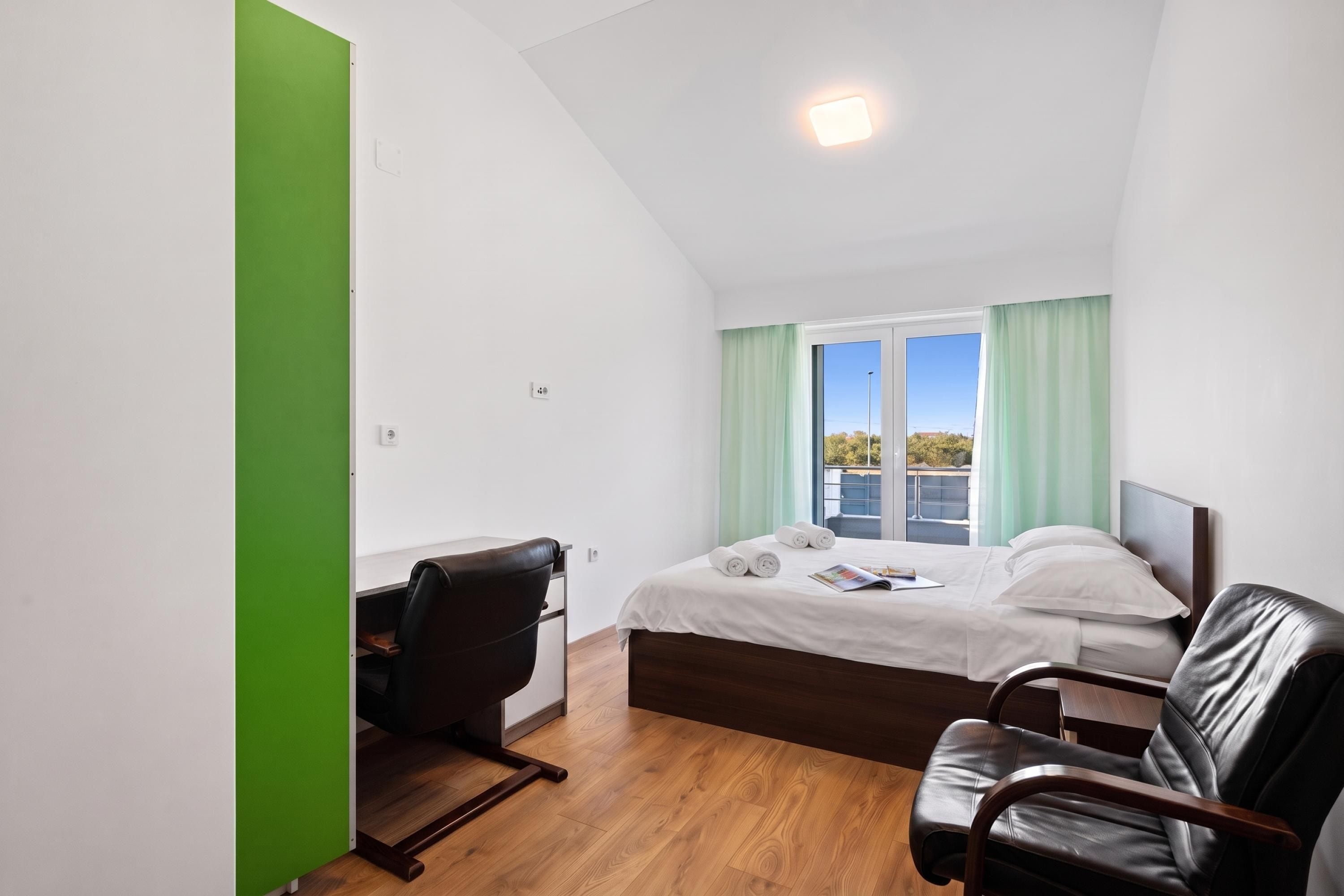 A modern hotel room with a bed, a desk, a chair, and a view of the outdoors.