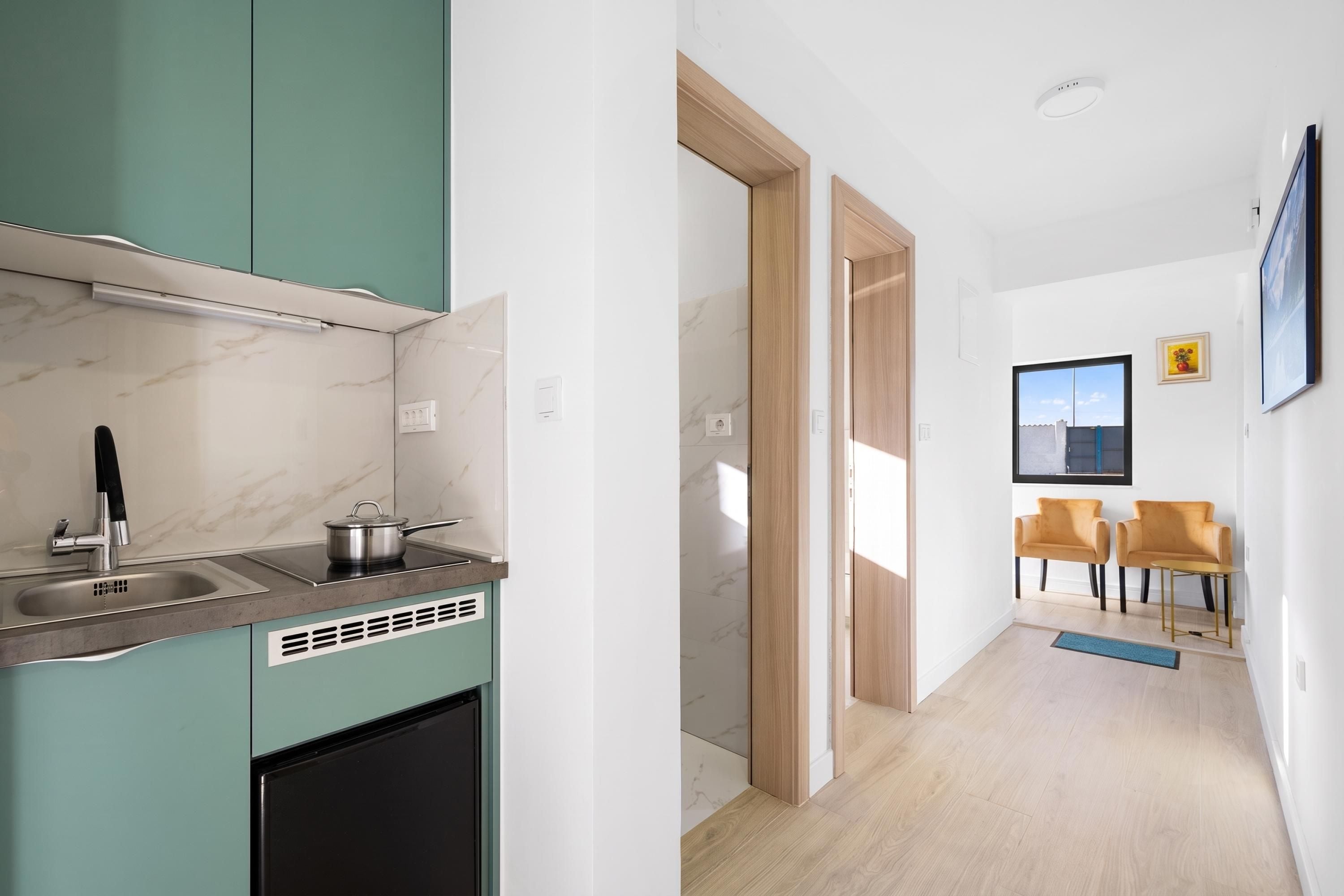 A modern kitchen with a built-in stove, sink, and cabinets. There is a hallway leading to a seating area with two chairs and a table.
