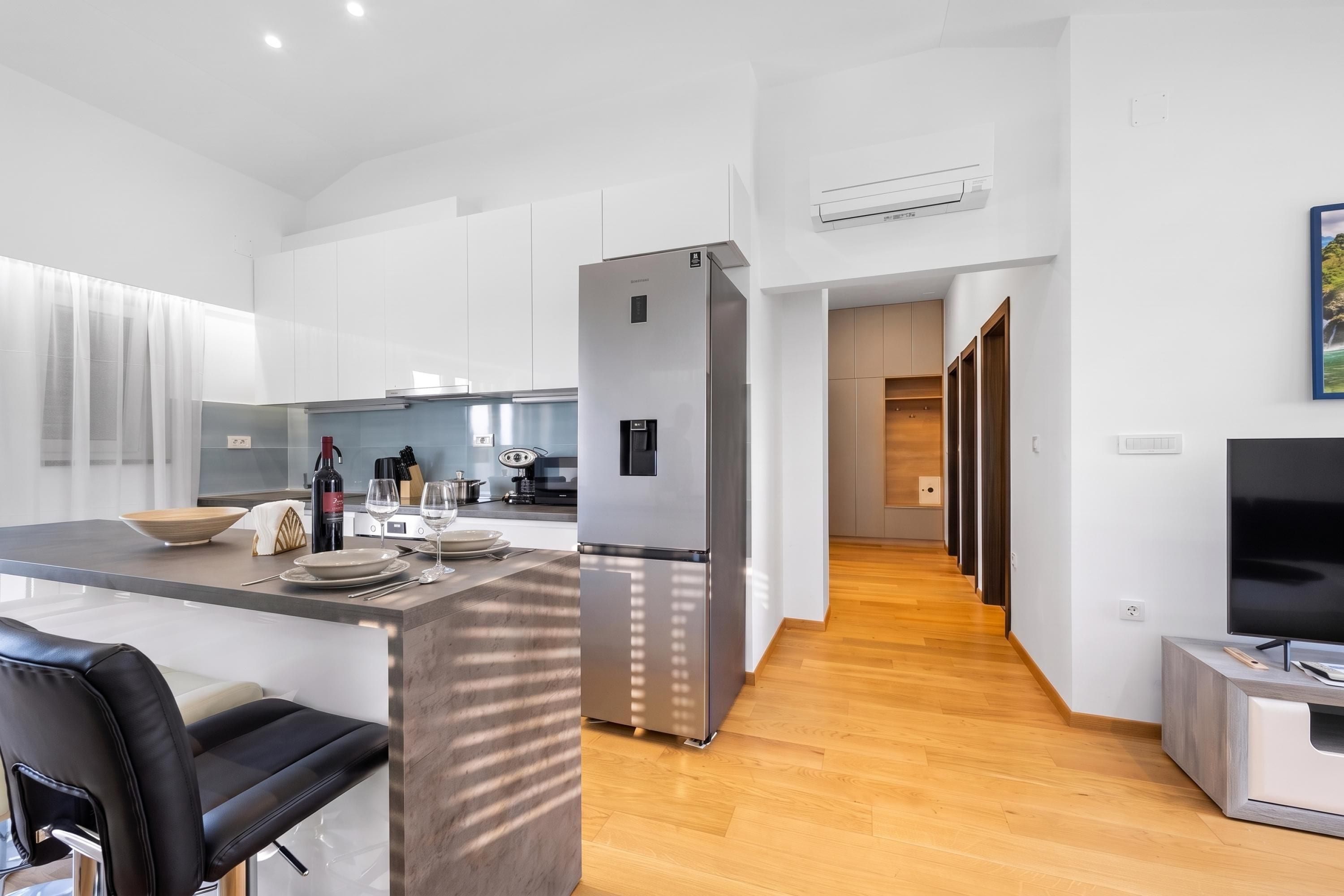 A modern kitchen with a large refrigerator, dining area with a table and chairs, and a hallway leading to a living area with a TV.