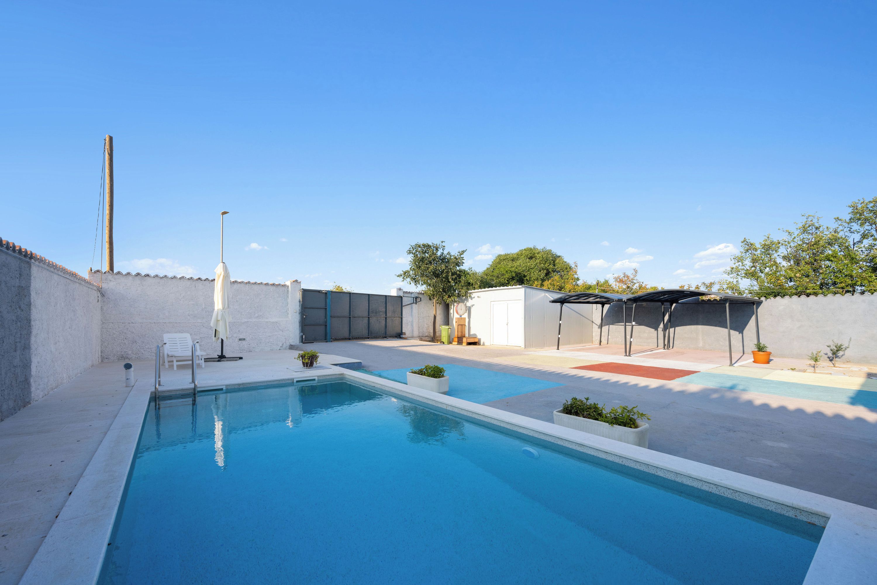 Una piscina amb aigua blava i cristal·lina, envoltada per un solàrium de formigó, una tanca blanca i algunes plantes en testos.