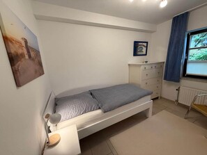 Ein Schlafzimmer mit einem Einzelbett, einem Nachttisch, einer Kommode, einem Fenster mit Vorhängen und einem gerahmten Zertifikat an der Wand.