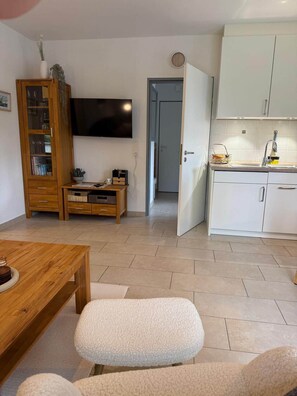 Ein modernes Wohnzimmer mit einem Holzespeisetisch, einem flachbildfernseher an der Wand, einer Küchenzeile mit weißen Schränken und einer Tür zu einem weiteren Raum.