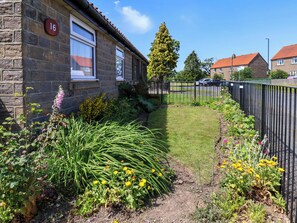 A stone building with a number '16' sign, a fenced garden with various plants, and a clear sky. - Wheeldale Cottage (Whitby)