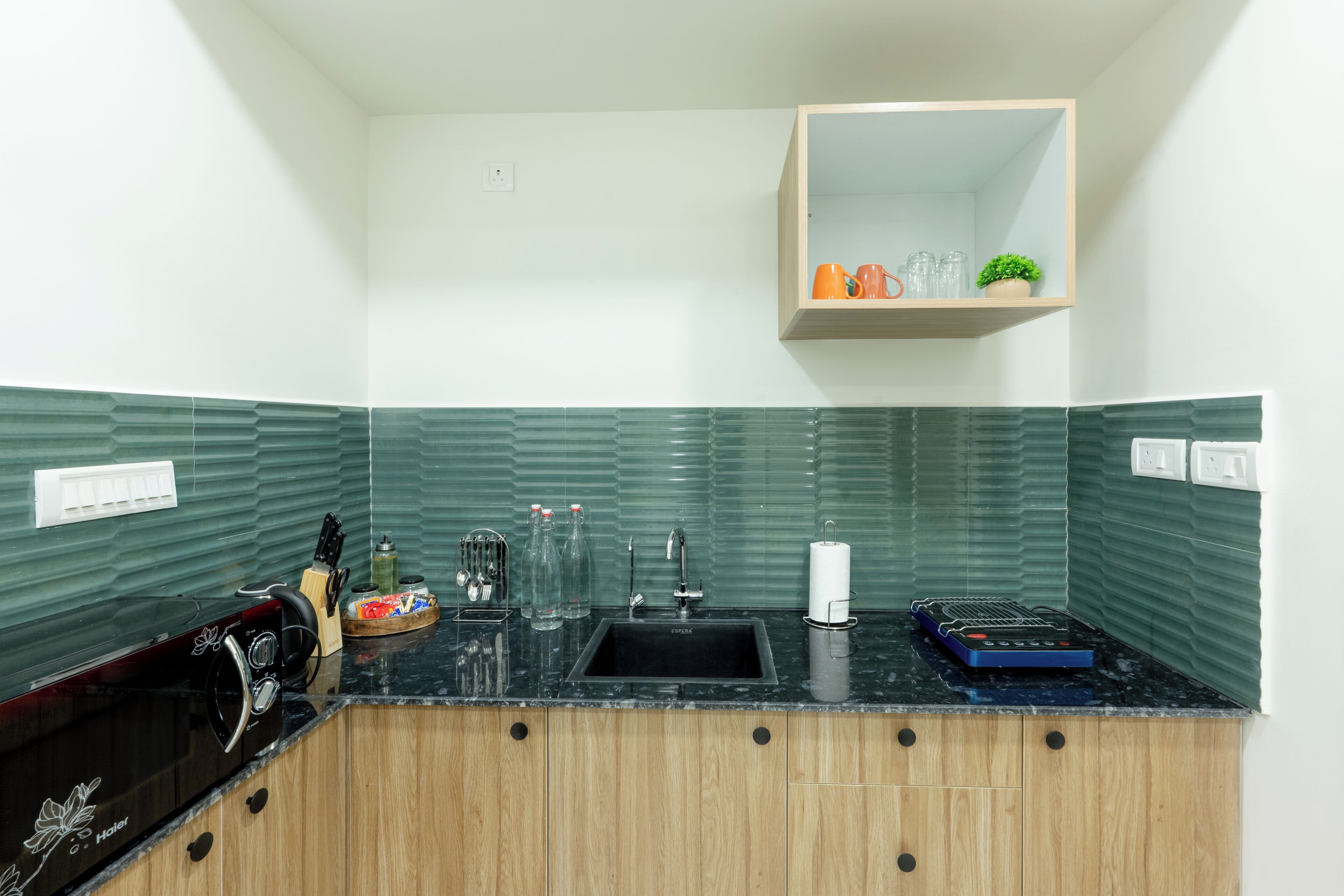 A kitchen with a black sink, wooden cabinets, a green tiled backsplash, and a small wooden shelf above the sink.