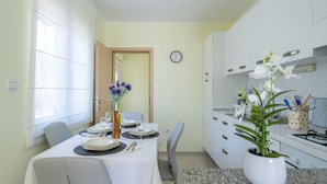 Un coin repas avec une table dressée pour quatre personnes, une horloge au mur et une cuisine dotée de meubles blancs et d’une cuisinière.