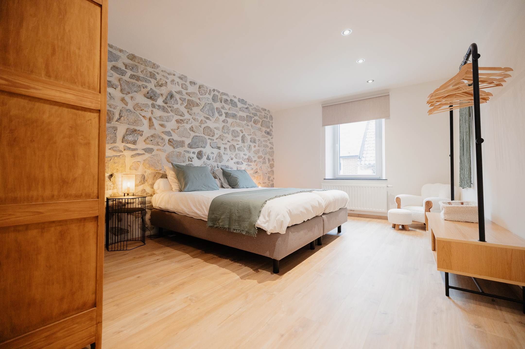 Un dormitorio con pared de piedra, cama con almohadas, mesita de noche, lámpara, ventana y una silla colgante.