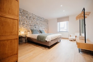 A bedroom with a stone wall, a bed with pillows, a bedside table, a lamp, a window, and a hanging chair. - 26 Pas de la Place (Gerpinnes)
