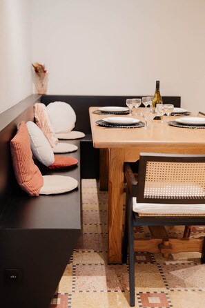 A dining area with a wooden table set for two, a bench with cushions, and a patterned floor. - 26 Pas de la Place (Gerpinnes)