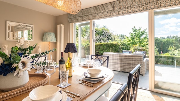 Un coin repas avec une table dressée pour quatre personnes, une grande fenêtre donnant sur un paysage verdoyant et un plafond décoré de motifs.
