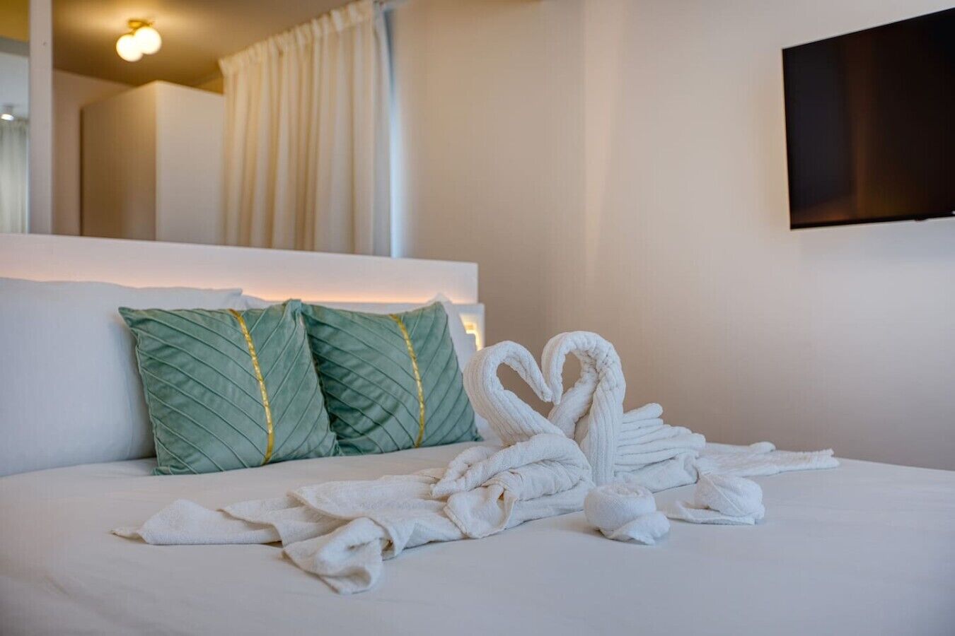 A hotel room with a neatly made bed, two green pillows, and a wall-mounted TV.
