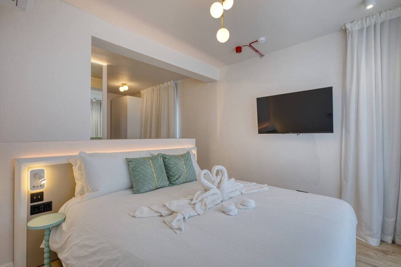 A modern hotel room with a large bed, a bedside table, a TV mounted on the wall, and a mirror above the bed.