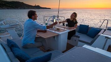 Un bateau avec un espace repas, une personne tenant un verre et une vue sur le coucher de soleil.