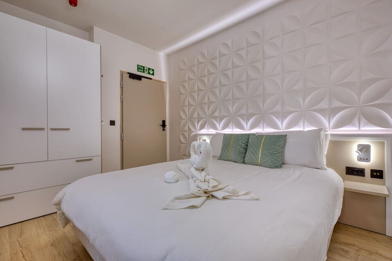 Ein Hotelzimmer mit Bett, Kleiderschrank, einer Tür mit Brandschutztürschild und einer Wand mit geometrischem Muster.