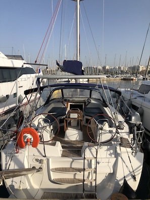A marina with multiple boats docked, a central boat with a wooden table and life preserver, and a clear sky. - Sun-Kissed Sailing Adventure in Islas Sarónicas (Alimos)
