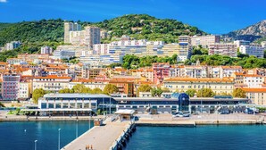 A coastal cityscape with a marina, buildings with varied architecture, and a hilly backdrop.