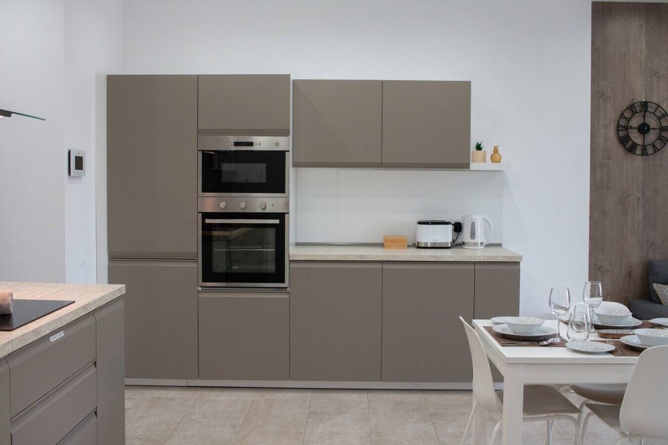 Een moderne keuken met inbouwapparatuur, een eethoek met een tafel gedekt voor twee personen, en een klok aan de muur.