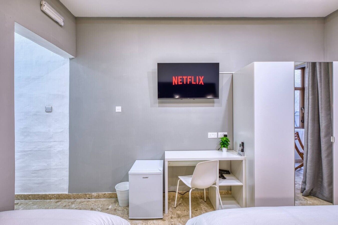 Een moderne hotelkamer met een bed, een bureau met stoel, een kleine koelkast, een aan de muur bevestigde televisie en een raam met gordijnen.