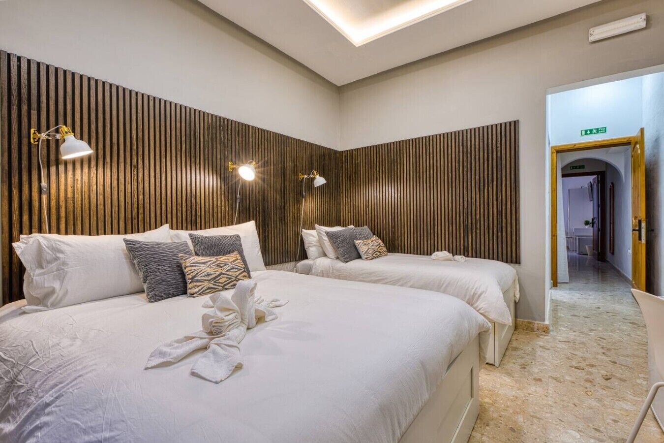 A hotel room with two beds, a wooden slat wall, and a hallway leading to an exit.