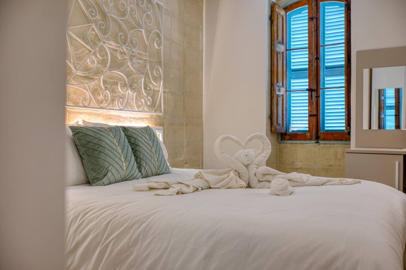 A bed with a heart-shaped towel arrangement, a decorative wall panel, and a window with shutters.