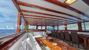 Interior d'un vaixell amb zona de menjador, seients i vista al mar.