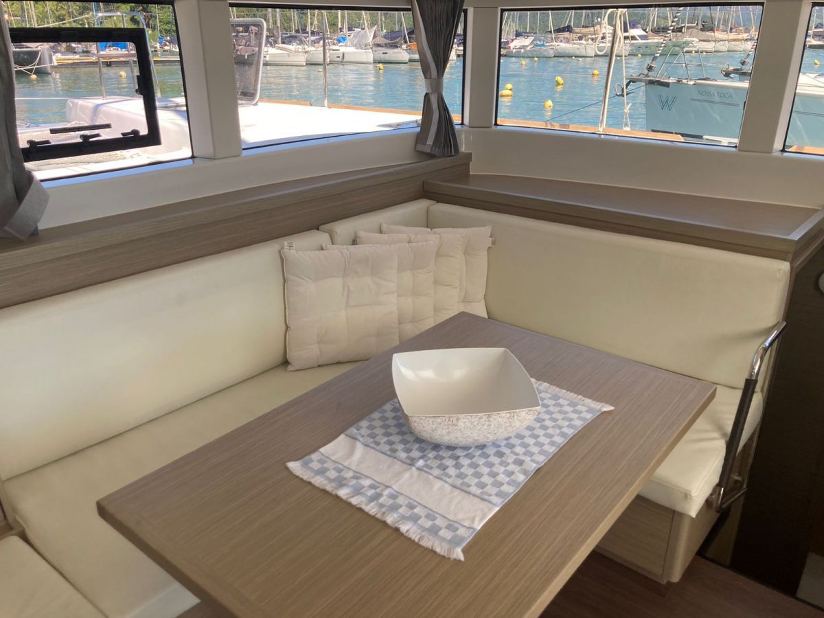 Interior de un barco con zona de asientos acolchada, una mesa con un bol y vistas a un puerto deportivo a través de amplias ventanas.