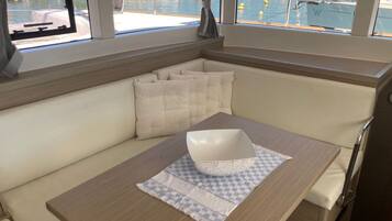 Interior de un barco con zona de asientos acolchada, una mesa con un bol y vistas a un puerto deportivo a través de amplias ventanas.