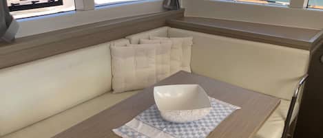 Interior de un barco con zona de asientos acolchada, una mesa con un bol y vistas a un puerto deportivo a través de amplias ventanas.