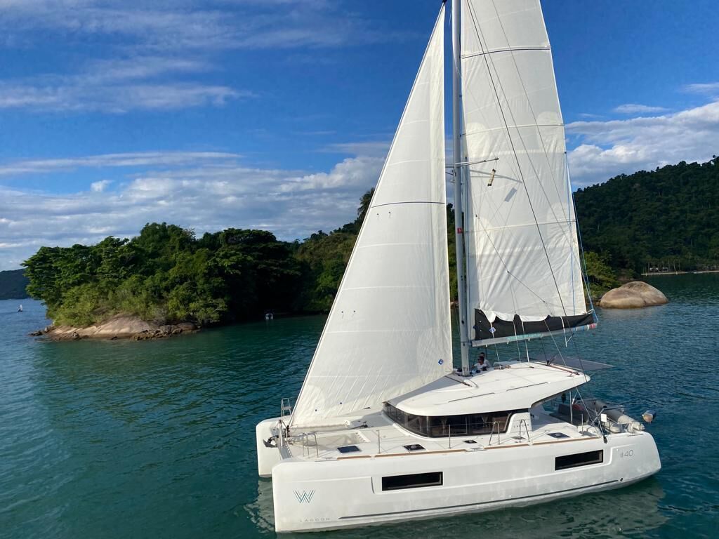 Un velero blanco con camarote y asientos en la cubierta, navegando sobre aguas tranquilas con una isla boscosa al fondo.