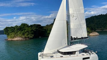 Un velero blanco con camarote y asientos en la cubierta, navegando sobre aguas tranquilas con una isla boscosa al fondo.