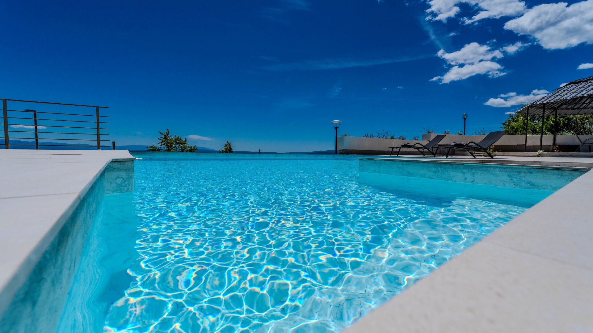A clear blue pool with a white deck, a metal railing, and a view of the ocean.
