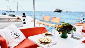 Schiffsinnenraum mit Esstisch, orangefarbenen Kissen, einem weißen Kissen mit einem Logo, einem Glas Wasser, einem Teller mit Essen und einer Vase mit Blumen.