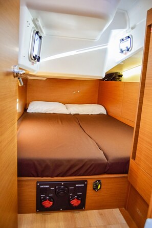 Interior compacto e bem iluminado, com uma cama de solteiro, um painel de controle para baterias e disjuntores, e um pequeno espaço de armazenamento.