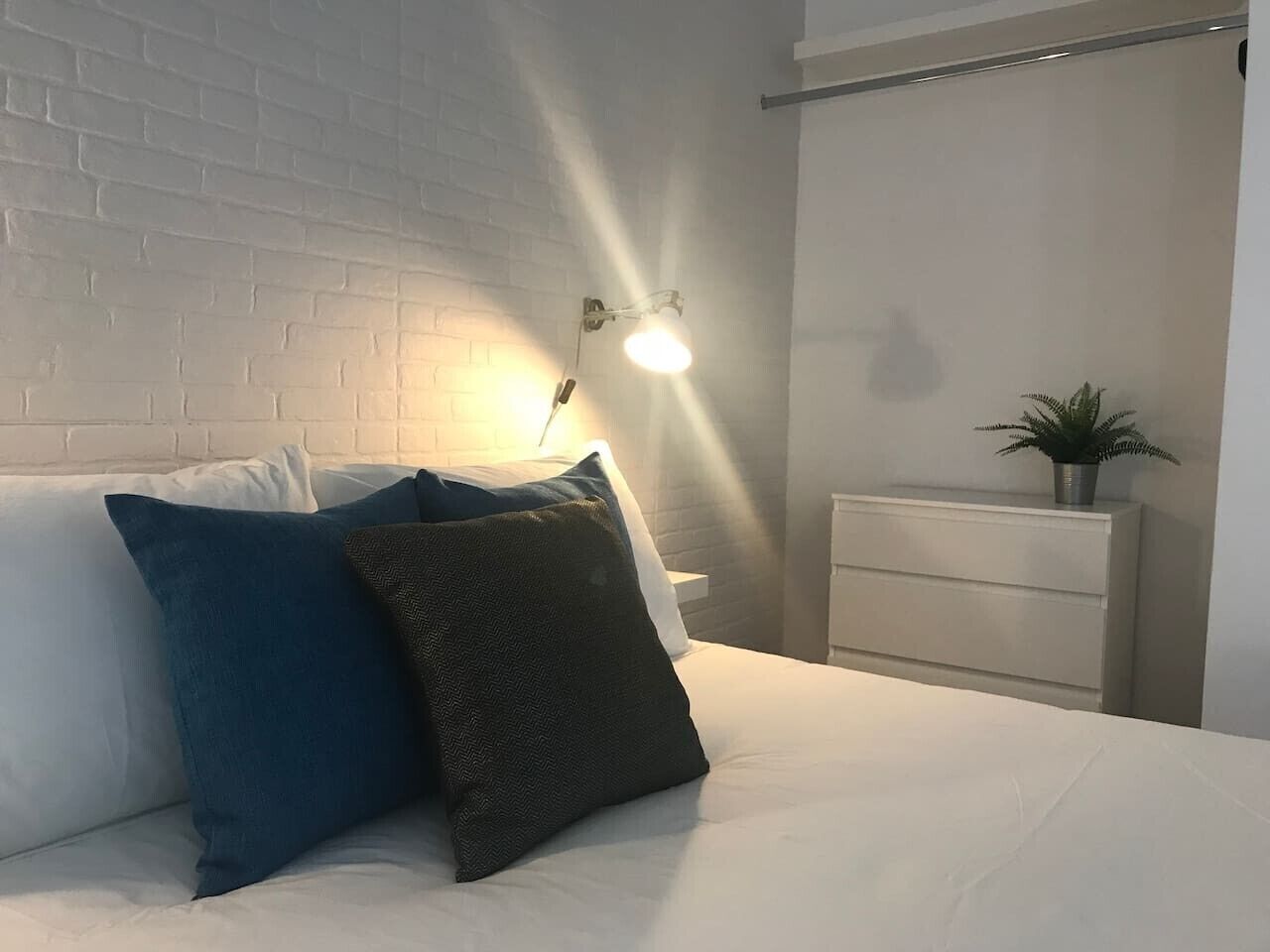 Une chambre avec un mur en briques blanches, un lit avec une literie blanche et deux oreillers, une table de chevet avec une lampe et une plante en pot.