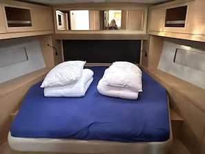 A narrow, single-bed room with a blue mattress, two pillows, and a headboard with a small window.