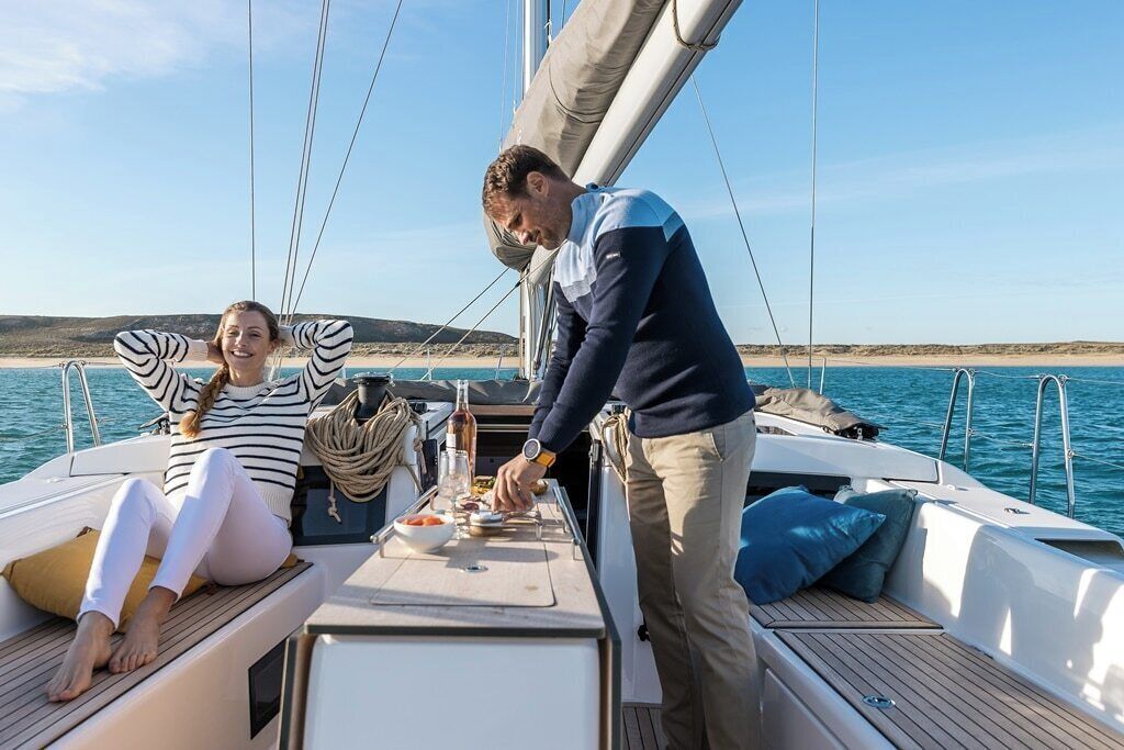 A sailboat with a woman and a man on deck, a table with food and drinks, and a clear view of the sea and distant land.