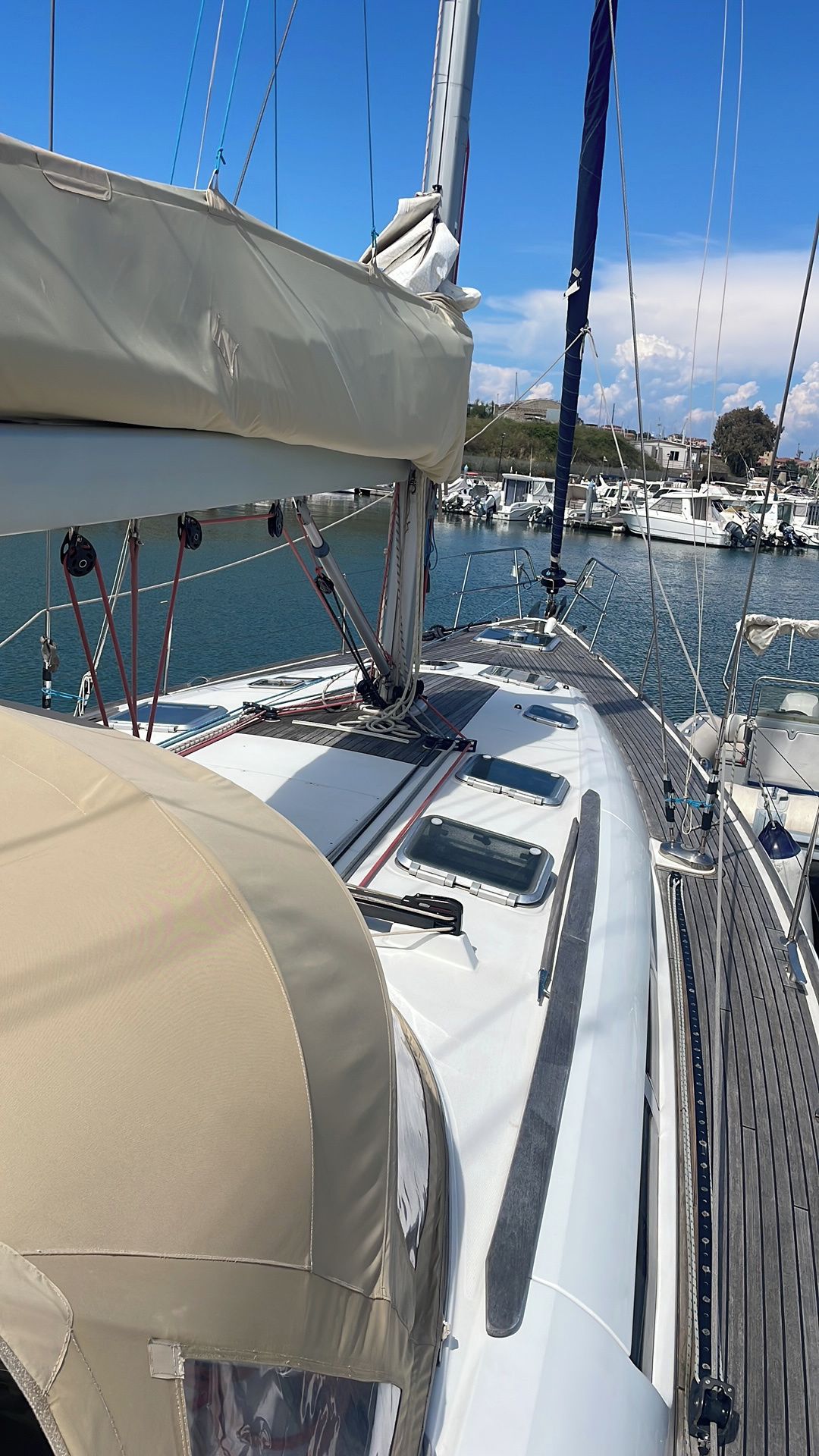 Het dek van een zeilboot met een afgedekte zone, een lier en een uitzicht op de jachthaven op de achtergrond.