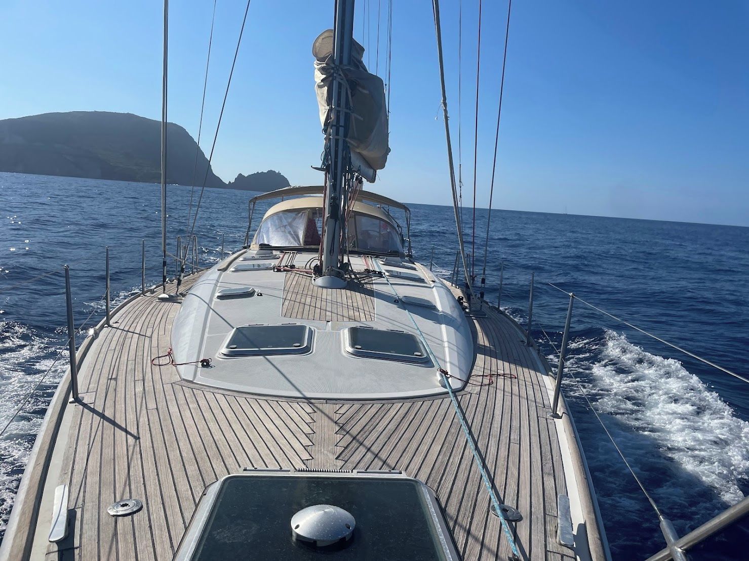 A sailboat's deck with a steering wheel, a mast, and a clear view of the sea and distant land.