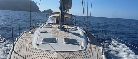 A sailboat's deck with a steering wheel, a mast, and a clear view of the sea and distant land.