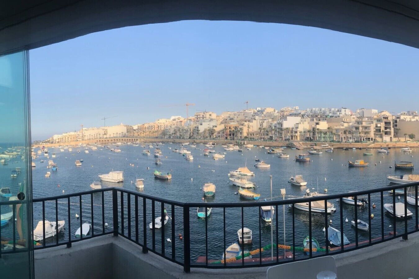 Een balkon met uitzicht op een haven vol boten, met een stadsbeeld op de achtergrond en een heldere lucht.