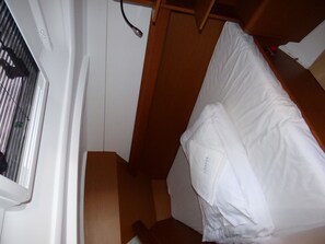 A narrow room with a bed, a window with blinds, and a wooden floor.