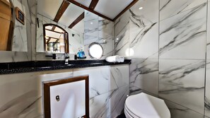 Une salle de bain avec des murs en marbre, un plan de toilette noir, un miroir, un miroir rond, des toilettes et une porte.