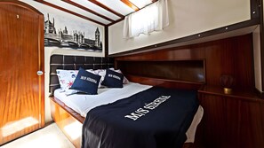 Une chambre à coucher avec un lit, une tête de lit, un mur orné d’un tableau représentant un paysage urbain, et une fenêtre avec des rideaux.